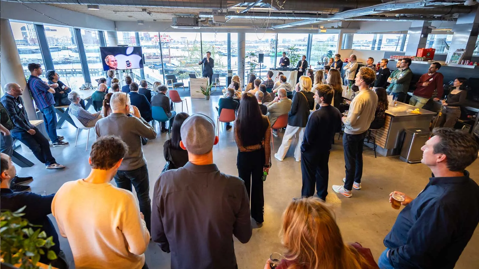 A large group of people stand in Apadmi's Amsterdam office with their backs to the camera, watching a presentation.