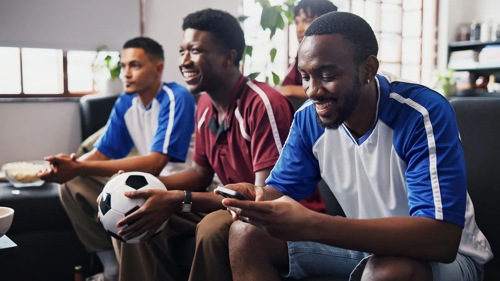Three men in sports jerseys relax in a bright living room, smiling while one checks a phone and another holds a soccer ball.
