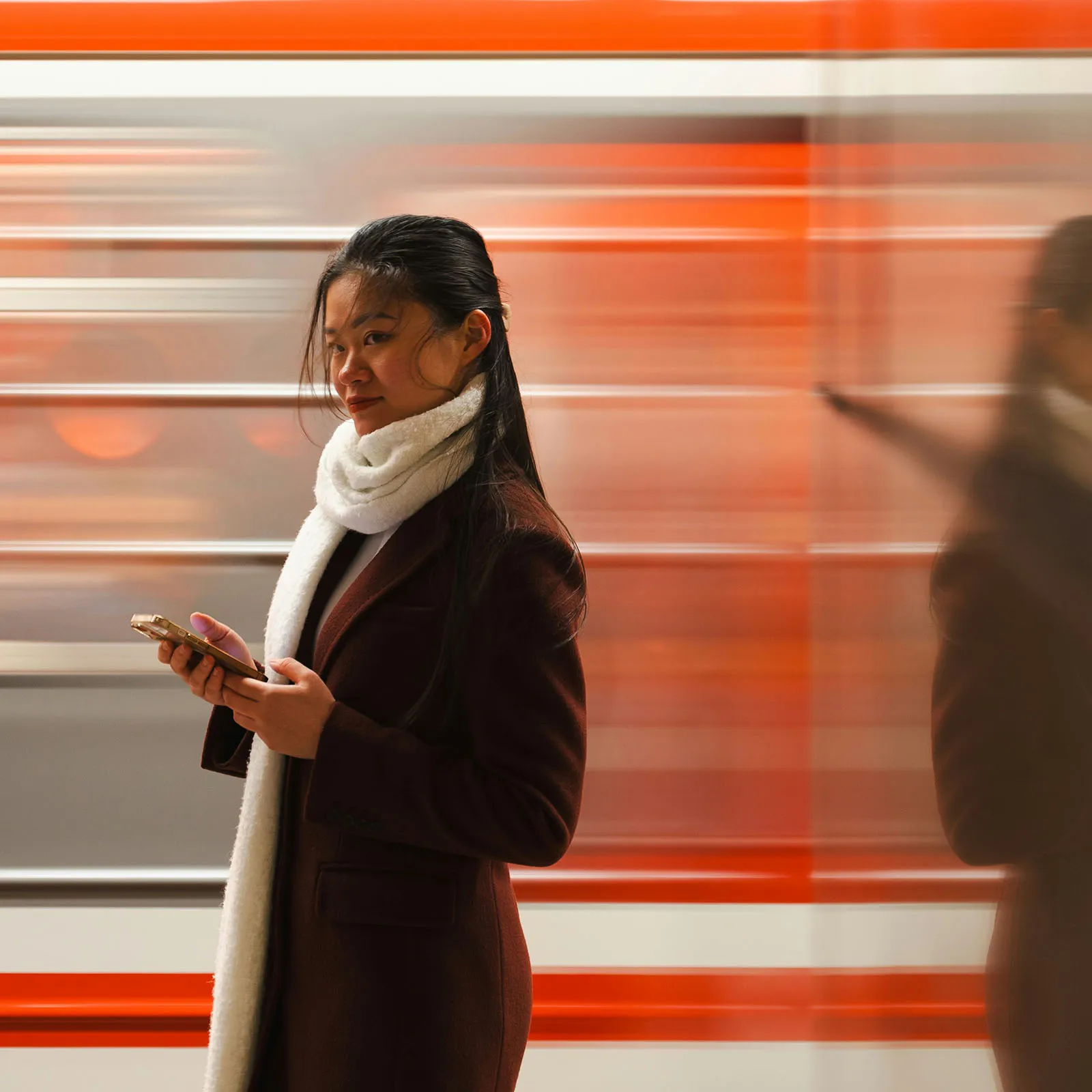 Young woman in a dark coat and white scarf checks her phone as an orange train rushes past.