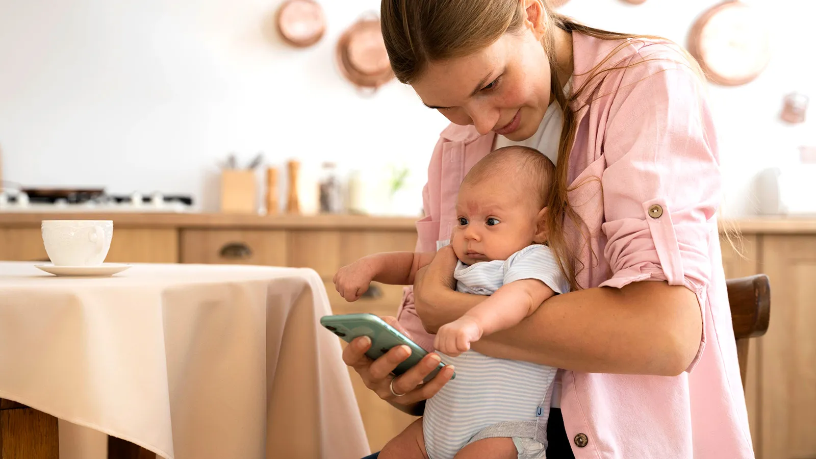 Mother in a pink shirt cradles her baby while checking a smartphone in a bright kitchen.