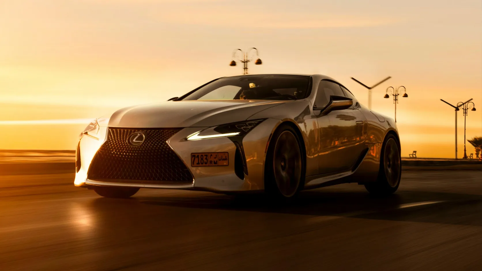 Silver Lexus coupe driving along a coastal road at sunset.