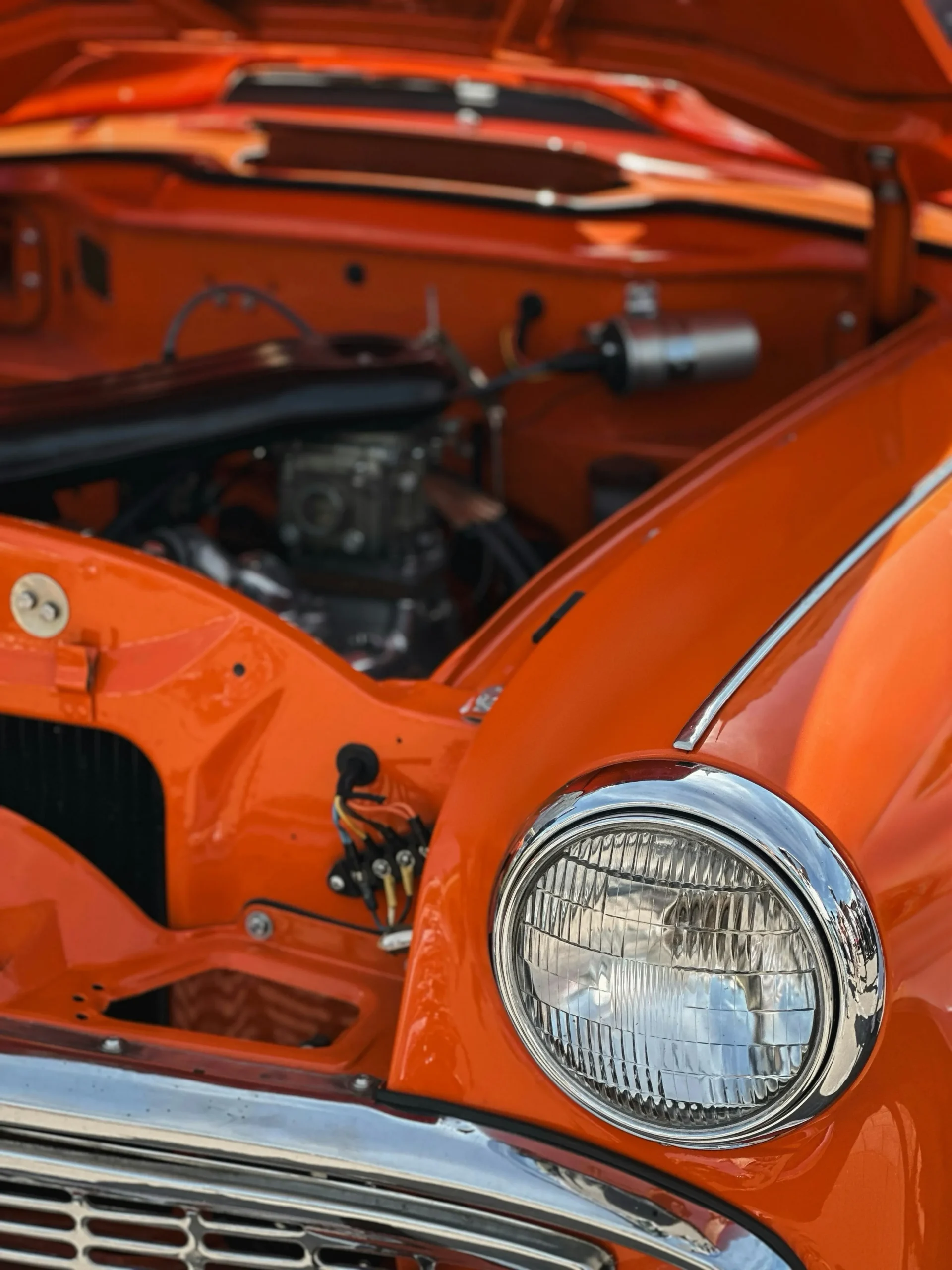 Close-up of an orange car with its hood open, exposed engine, and large round chrome headlight.