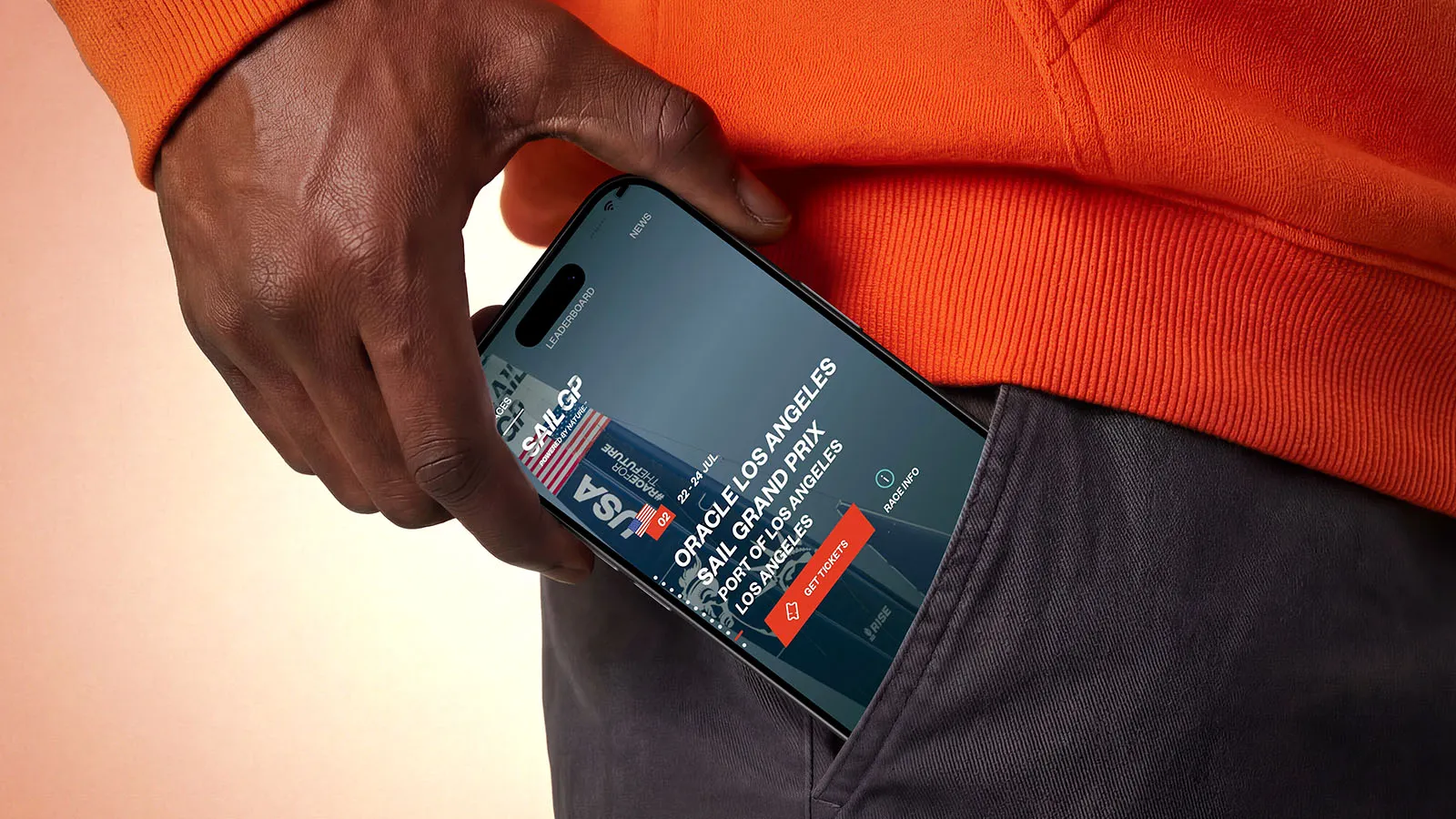 Close-up of a hand holding a smartphone displaying a SailGP Los Angeles event listing, partly tucked into dark trousers.