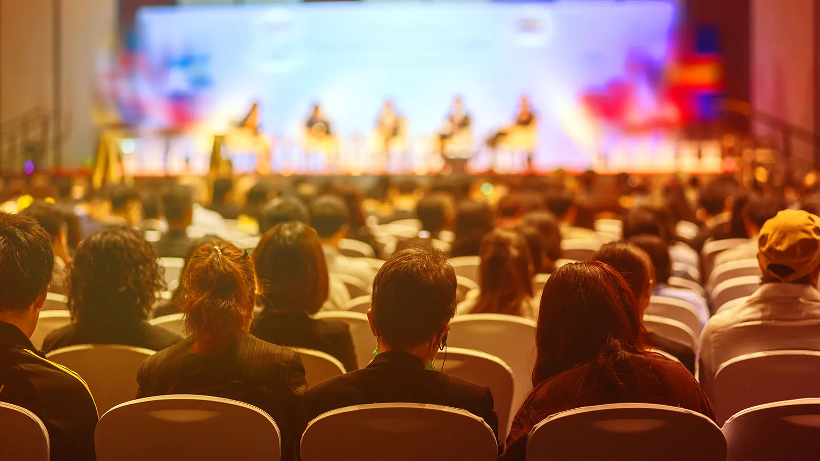 Blurred image showing a corporate event, with an audience looking towards a panel of people talking.