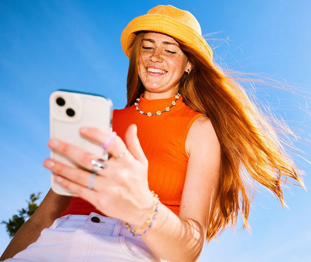 Smiling young woman in a yellow sunhat looks at her smartphone outdoors; her long red hair blows in the sunlight against a blue sky.