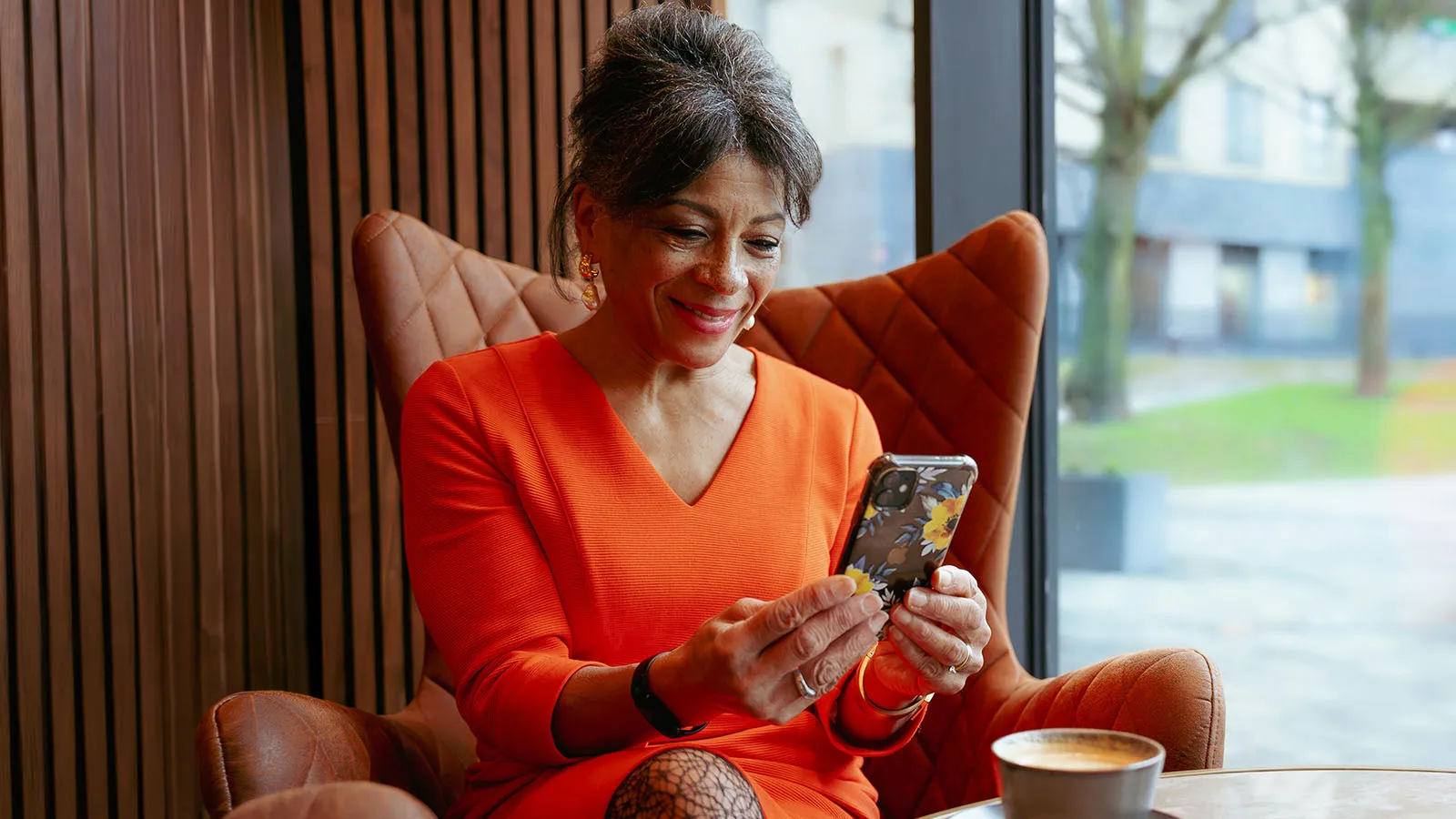 Smiling woman in an orange sweater sits in a leather armchair, holding a decorated smartphone beside a coffee cup in a bright café.