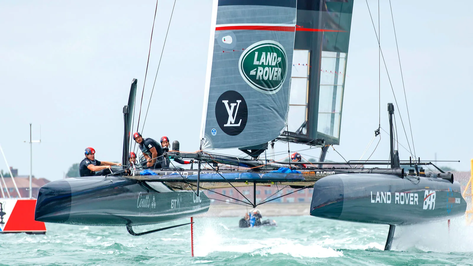Land Rover-branded foiling catamaran races across choppy water, crew members aboard beneath a large sail in a fast-paced sailing scene.