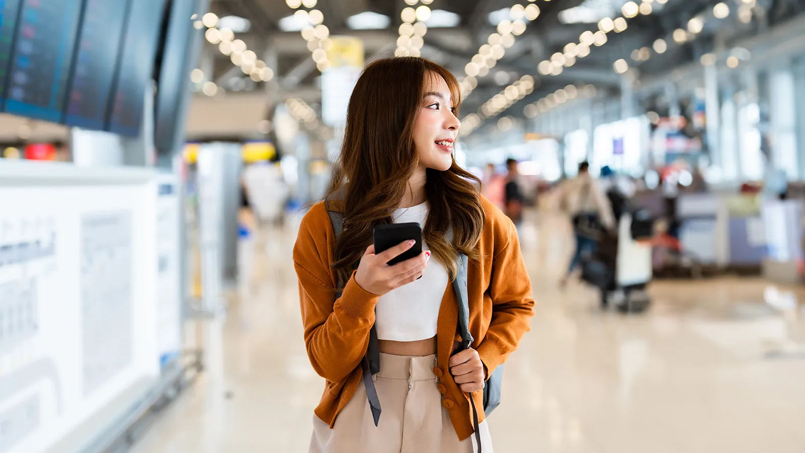 Smiling young woman in an airport terminal holds a smartphone and backpack strap, wearing a brown jacket as travelers blur behind her.